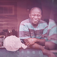 Homem sorrindo apoiado na janela de casa, usando camiseta listrada